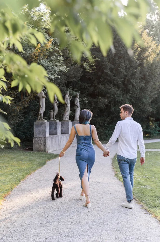 Couple walks hand-in-hand with a small dog on a gravel path in a park, surrounded by greenery and classical statues in the background.