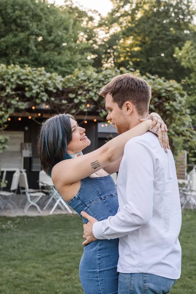 Couple embracing outdoors; woman with blue hair in denim dress, man in white shirt. Grassy area with blurred background.