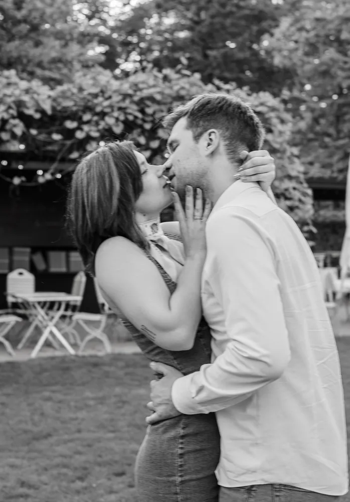 Couple sharing a kiss in a garden, woman in sleeveless dress, man in white shirt, affectionate embrace, outdoor setting with chairs and trees in the background.