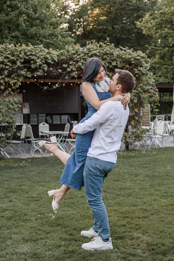 Couple dancing outdoors, man in white shirt and jeans lifts woman in denim dress, barefoot, grassy area with patio furniture and trees in background.