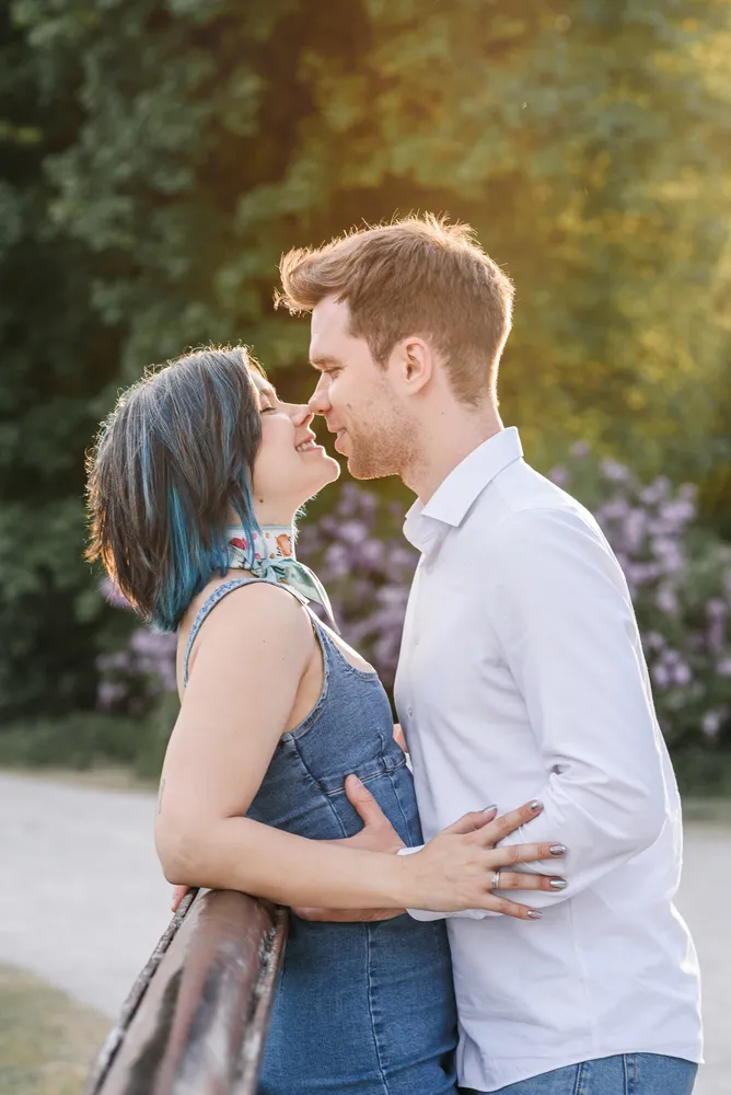A couple shares a kiss in a park, she with blue-tinted hair, he in a white shirt, both dressed casually, surrounded by greenery and purple flowers.