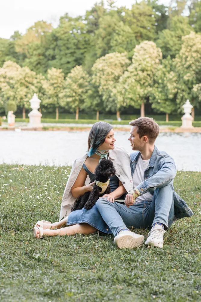 Couple and dog sitting on grass in park. Woman with blue hair, man in jeans. Both smiling, facing each other. Water and trees in background.
