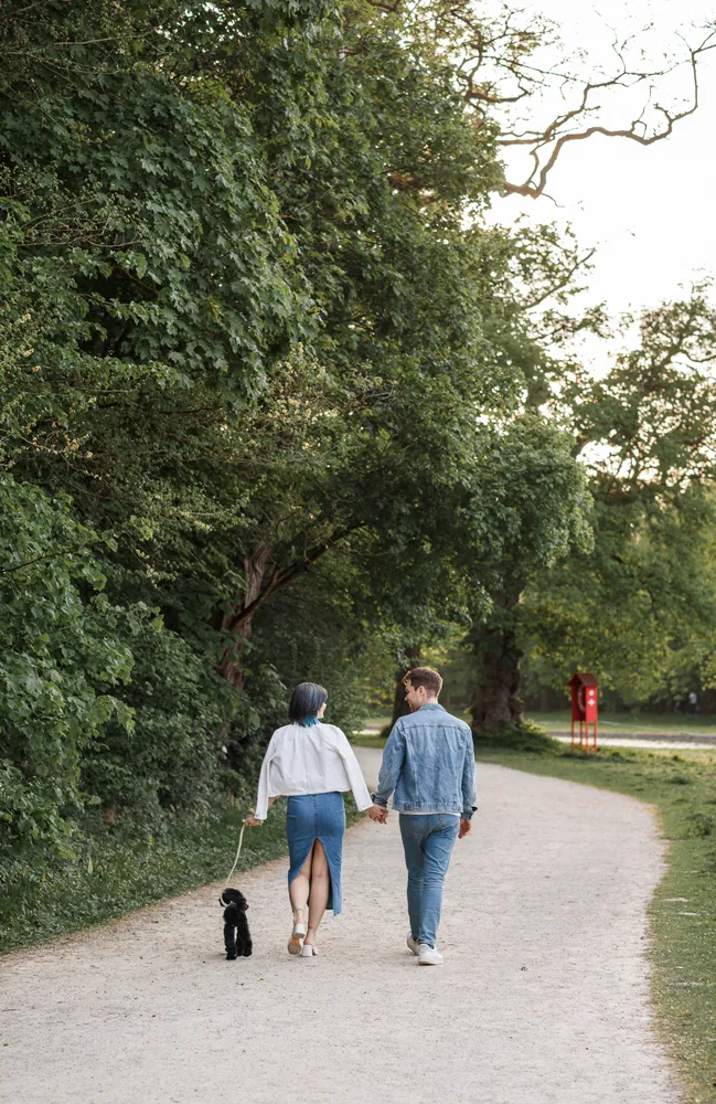 A couple strolls on a gravel path, woman in white blouse and blue skirt leading a black dog, man in denim jacket and jeans by her side. Trees and grass frame the scene.