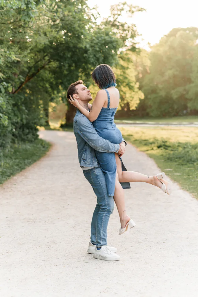 A man in a denim jacket and jeans lifts a woman in a denim dress on a gravel path surrounded by trees. They embrace, smiling, in the soft glow of daylight.