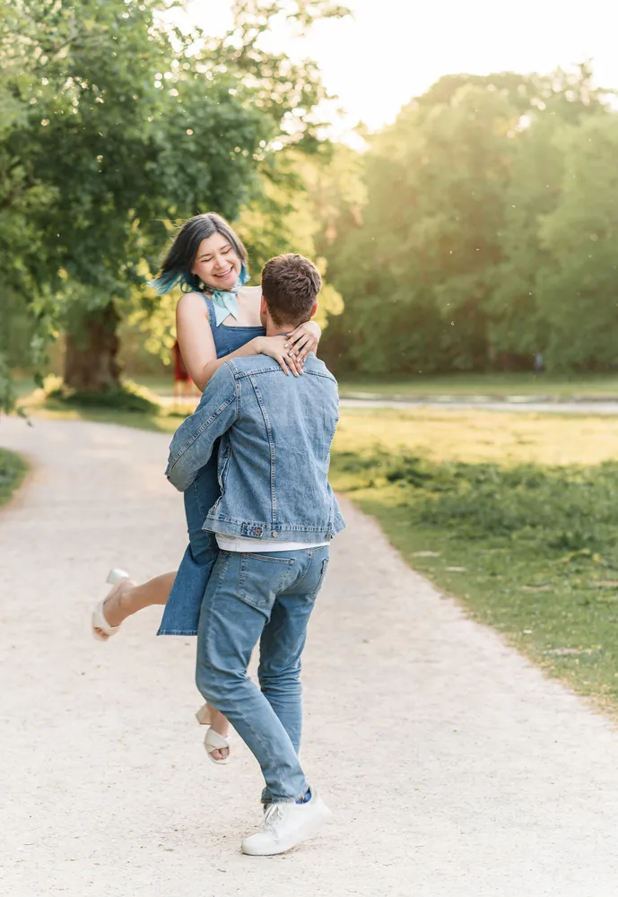 Couple in casual attire shares joyful embrace on sunlit park path, woman's blue hair contrasting with greenery.