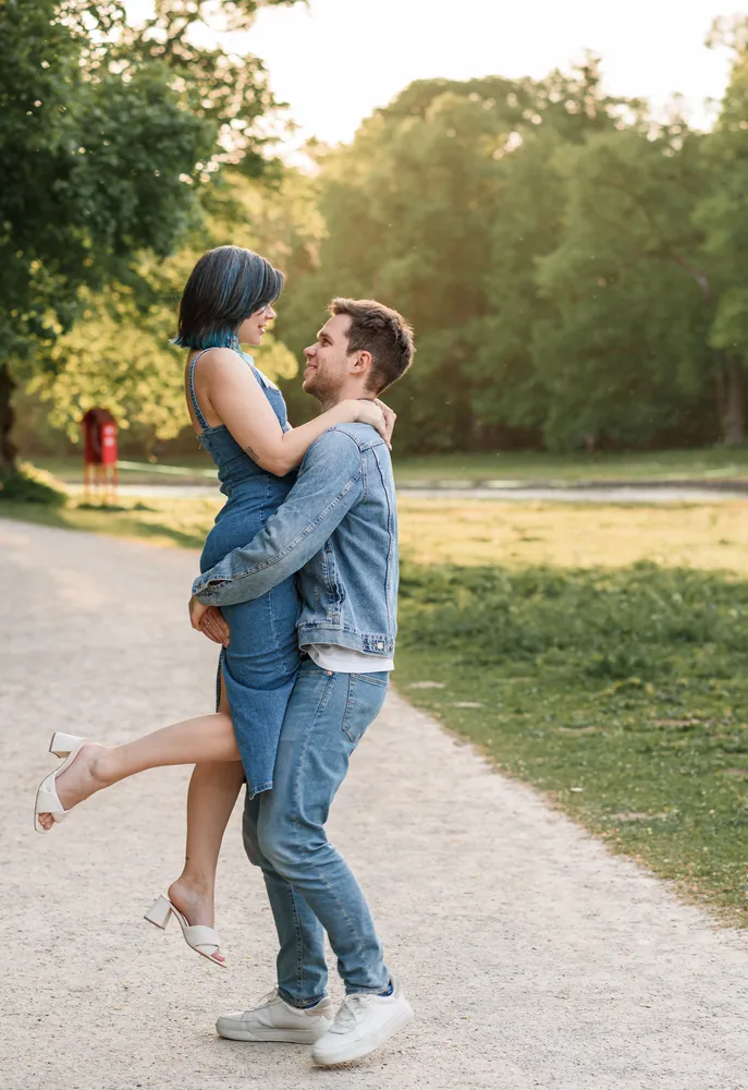A couple shares a joyful dance in a park; he lifts her in a denim-clad embrace on a sunlit path, surrounded by greenery.