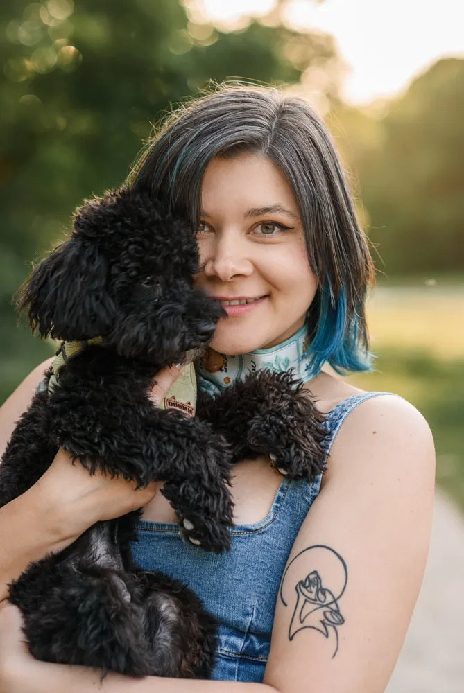 A young woman with blue-tipped hair smiles while holding a black poodle in a park. She wears a denim tank top and has a wolf tattoo on her arm.