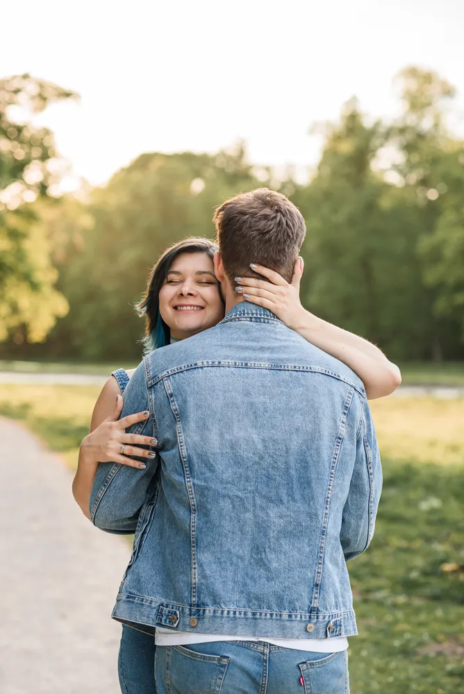 A couple embracing on a park path, both wearing denim jackets. The woman's face is visible, smiling, while the man's face is not shown. The background is a blurred natural landscape.
