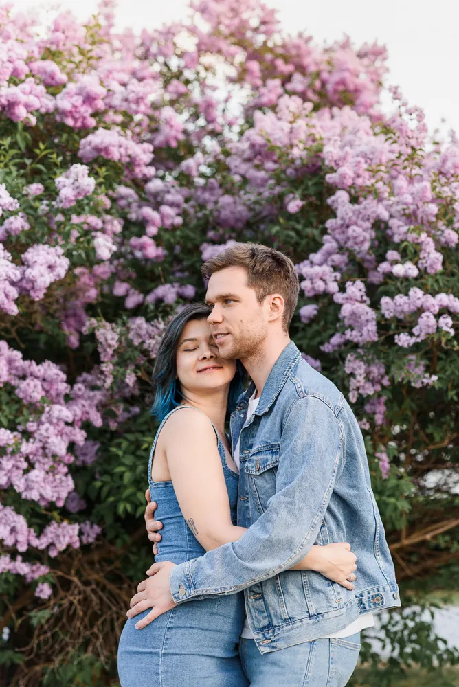 Couple embracing in front of vibrant lilac bush, woman with blue-tinted hair, both in denim attire, tender moment captured outdoors.