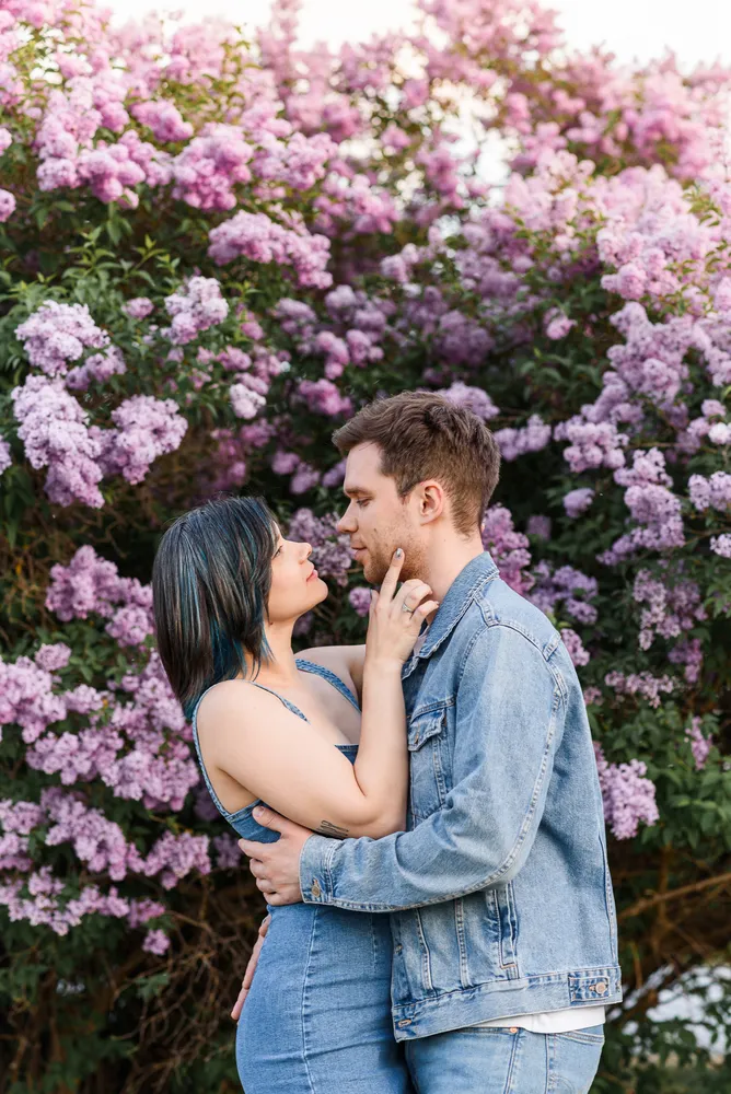 Couple in denim embrace before lilac blossoms, her hand tenderly touches his face.