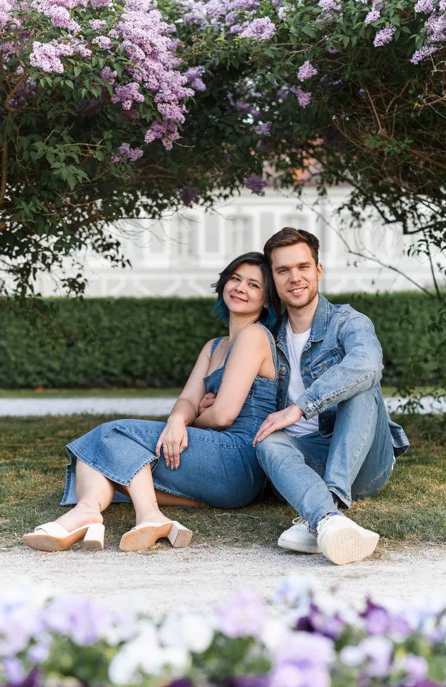 A couple in denim attire smiles outdoors, seated under a lilac tree, with a white building in the background.
