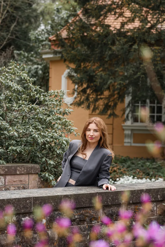 Woman in grey blazer leans on stone wall, gazing at camera. Behind her, a yellow house with white trim is partially obscured by trees and shrubs. Vibrant purple flowers in foreground.