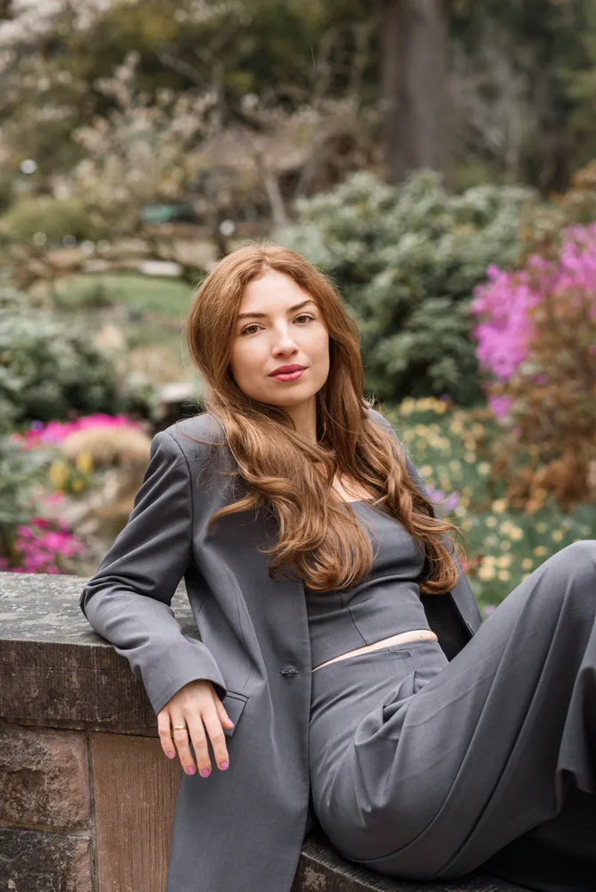 Woman in grey suit leans on stone wall in garden, manicured nails, natural makeup, soft focus background.