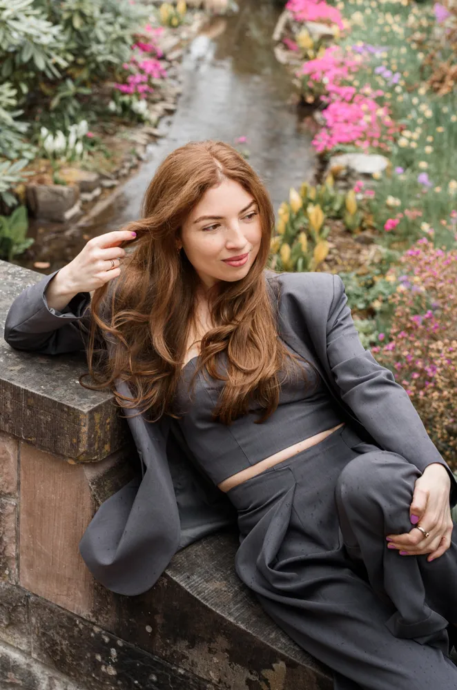Woman in gray suit leans on stone wall in garden, gazing thoughtfully at camera, with colorful flowers and a stream in the background.