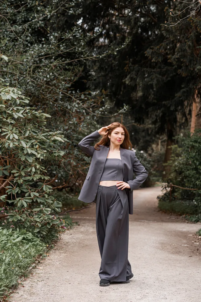 Woman in grey suit poses on a forest path, hand on head, surrounded by greenery.