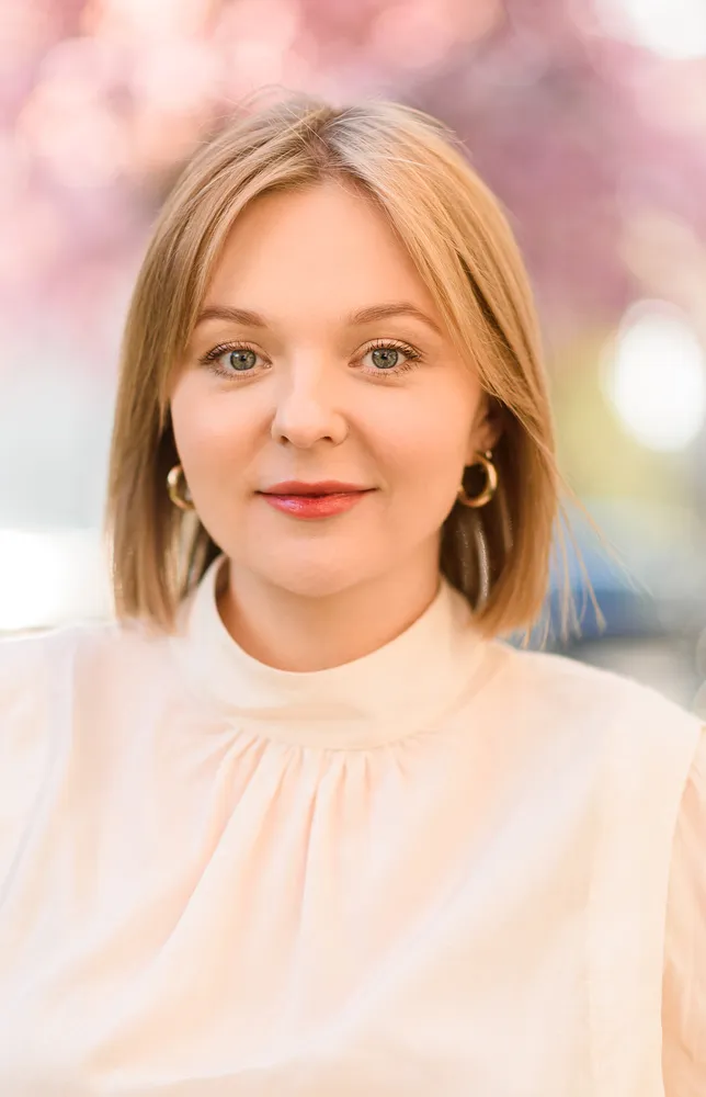 Woman in white blouse, gold earrings, soft smile, blurred pink background.