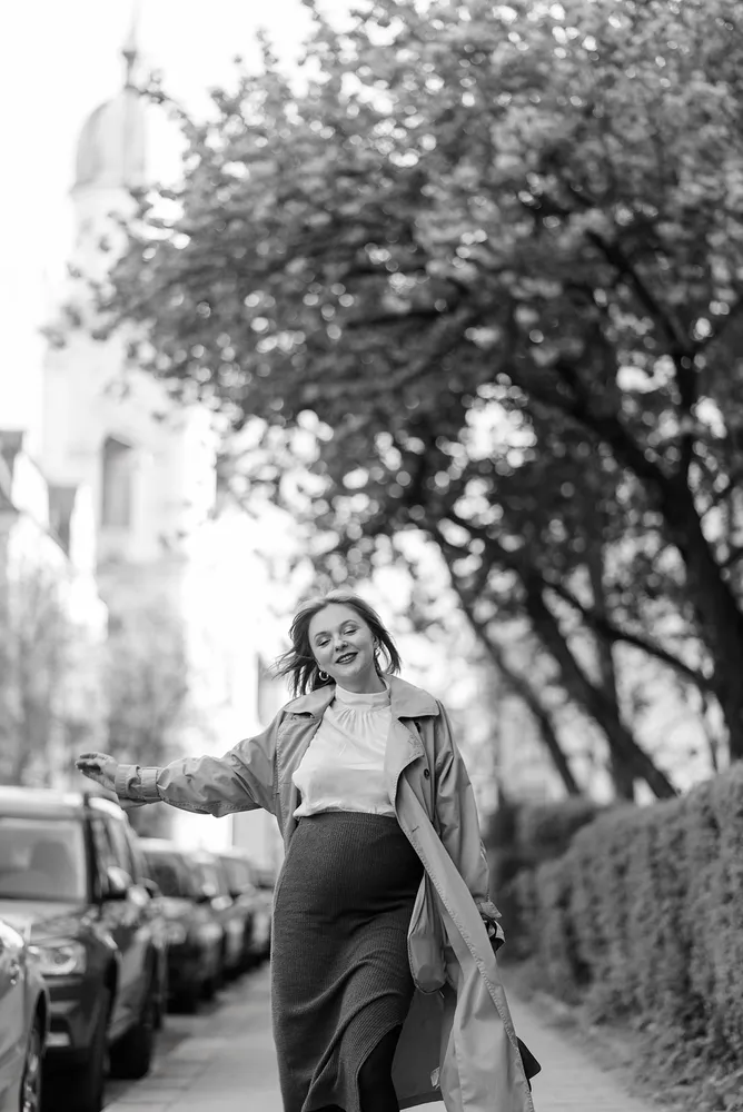 Woman in light coat and skirt walks down tree-lined street, smiling, cars parked on side, old building in background.