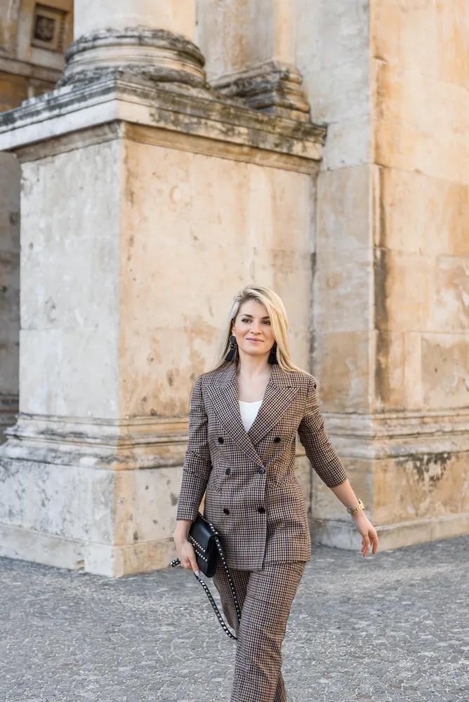 Woman in brown plaid suit with white top walks past ancient stone building with columns, holding black handbag, wearing gold watch.