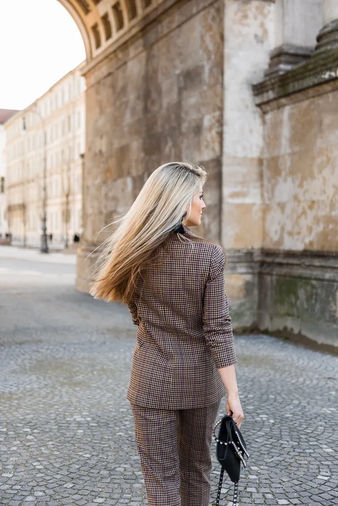 Woman with long blonde hair walks down cobblestone street, wearing a plaid blazer and matching pants, carrying a black purse, in front of a historic stone building with an arched entrance.