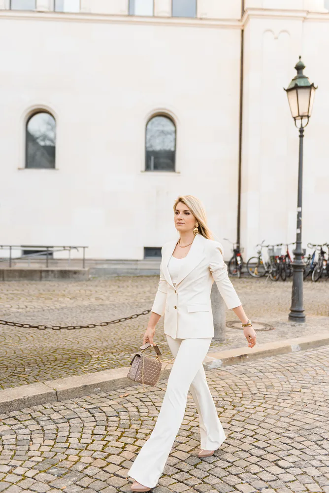 Woman in white suit walks on cobblestone street, holding brown bag, past a white building with arched windows and parked bicycles.