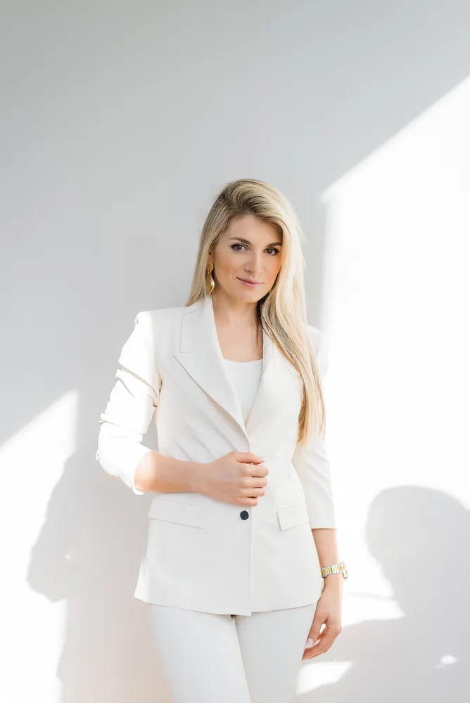 Woman in white suit stands against a plain background, hands clasped at waist, professional portrait.